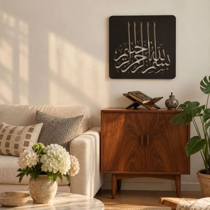 Bismillahirrahmanirrahim yazılı siyah metal Besmele tablosunun modern salonda, açık renkli koltuk üzerinde konumlandırılmış hâli; Kufi hat sanatı tasarımıyla sade ve çağdaş İslami duvar dekorasyonu.