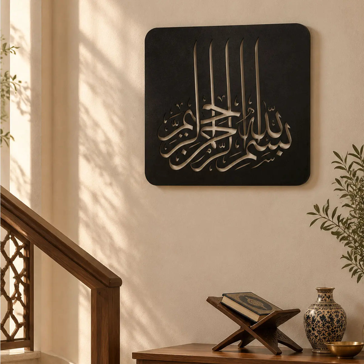 Modern antre ve giriş alanında duvara asılmış siyah metal Besmele tablosu; Bismillahirrahmanirrahim yazısının Kufi hat sanatıyla tasarlandığı İslami duvar dekoru, açık renk duvar ve bitki detaylarıyla tamamlanıyor.